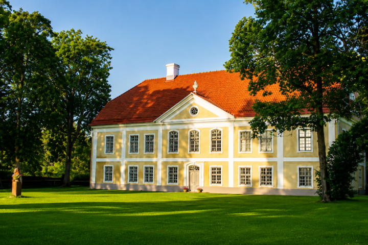 Yellow mansion with red roof, surrounded by green trees and grass.