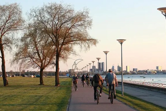 People cycling on a path along a waterfront with trees and streetlights.