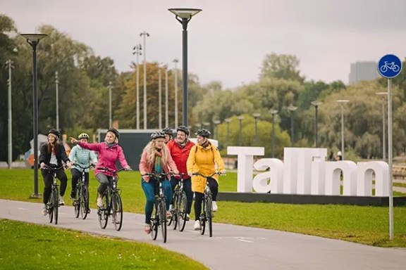 Group of cyclists riding on a path near large Tallinn sign.